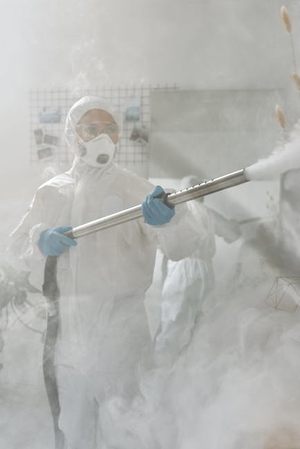 A professional cleaner from Colliers Wood Cleaners is performing deep surface cleaning in an indoor space, wearing protective clothing including a white coverall, face mask, goggles, and blue gloves. 