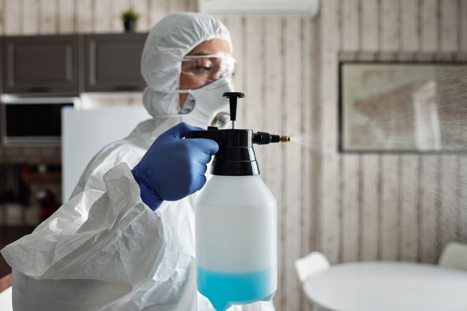 A professional cleaner from Colliers Wood Cleaners is performing deep surface cleaning in an indoor space, wearing protective clothing including a white coverall, face mask, goggles, and blue gloves. 