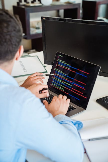 A person seated at a desk working on a laptop displaying colorful lines of code, with a second monitor placed in front. The desk contains office supplies, including a notebook and a remote control, an
